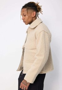 Veste en laine beige avec un col, des poches avant et une fermeture éclair. Caractérisée par un tissu texturé et une coupe décontractée.