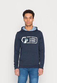 Felpa blu navy con una fodera della cappuccio di un blu chiaro. Presenta una grande grafica bianca "JACK & JONES", cordini regolabili e una tasca frontale.