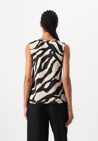 Personne aux cheveux tressés portant un haut sans manches à imprimé zèbre noir et beige, et un pantalon large noir, tournant le dos à la caméra.