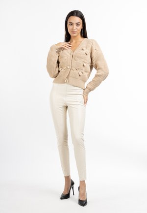 Cardigan lavorato a maglia beige con grandi bottoni e dettagli testurizzati, abbinato a pantaloni aderenti color crema e tacchi alti neri.