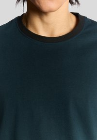 Mørk teal t-shirt med en sort ribbet rund halsudskæring, glat tekstur og korte ærmer, med et enkelt, udekoreret design.