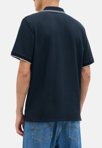 Polo shirt bleu marine avec un motif texturé, bord blanc au col et aux manches, et fentes sur les côtés. Assorti avec un jean bleu clair.