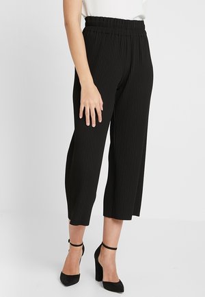 Kobieta w czarnych szerokich spodniach o długości do połowy łydki z elastycznym pasem i czarnych sandałach na słupku z paskiem wokół kostki, stojąca na jednolitym tle.