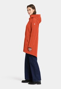 Veste imperméable orange avec capuche, fermeture éclair frontale, deux poches latérales, et coupe décontractée, associée à un jean bleu foncé à jambes larges et des bottes noires.
