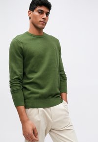 Πράσινο υφαντό πουλόβερ με στρογγυλή λαιμόκοψη και ριμπ μανσέτες, με ένα μικρό λογότυπο στο στήθος. Συνδυασμένο με ανοιχτό μπεζ παντελόνι.