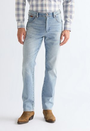 Mand iført lyseblå jeans, brune ruskindsstøvler og en hvid og blå ternet skjorte, der er stoppet ned, vist fra taljen til fødderne.