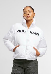 Bijela puffer jakna s visokim ovratnikom, crnim logotipom na prsima i elastičnim manžetama. Uključeni su džepovi radi funkcionalnosti.