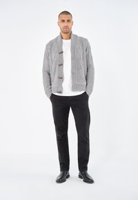 Grå stickad cardigan med kabelmönster, träknappar och sjalkrage, bärs över en vit T-shirt och i kombination med svarta byxor.
