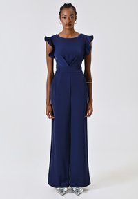 Navyblauer Overall mit Rüschenshortärmeln, tailliertem Oberteil, weitem Bein und einem v-förmigen Detail an der Vorderseite. Getragen mit metallischen spitzen Schuhen.