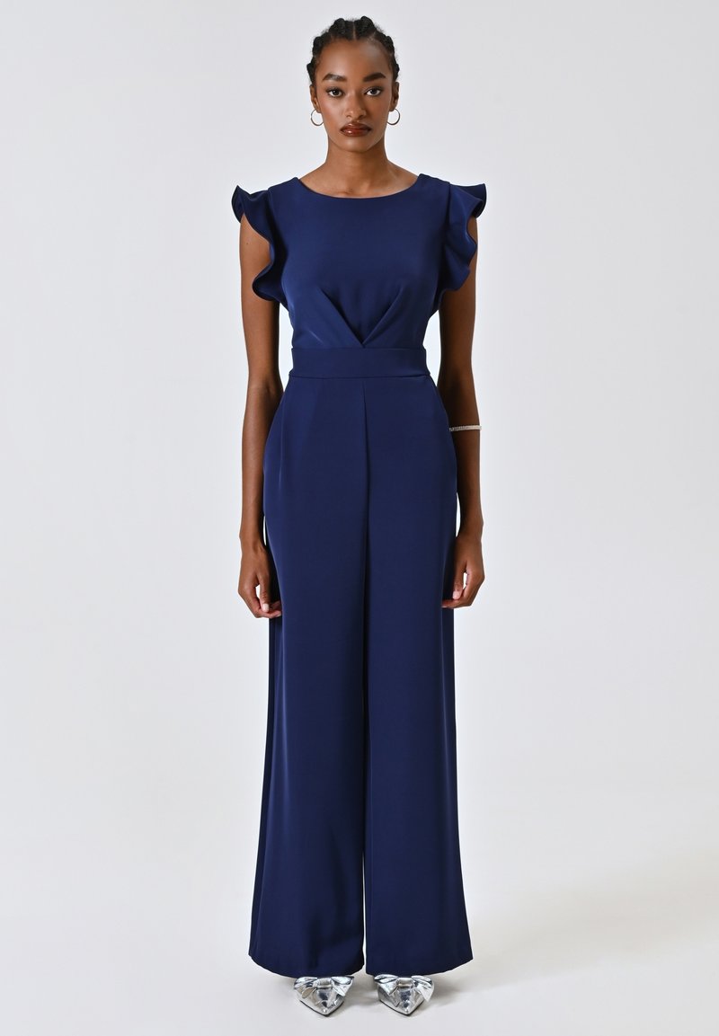 Navyblauer Overall mit Rüschenshortärmeln, tailliertem Oberteil, weitem Bein und einem v-förmigen Detail an der Vorderseite. Getragen mit metallischen spitzen Schuhen.