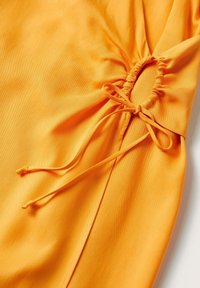 Tissu orange vif avec un détail froncé rassemblé noué en un nœud à l'aide de cordons orange assortis.