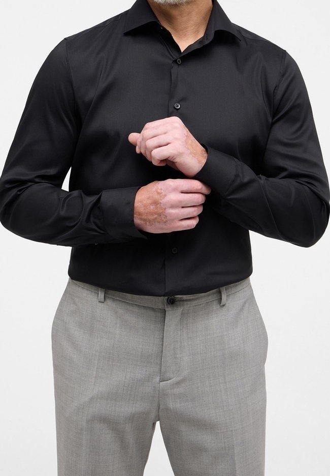 Homme ajustant la manche d'une chemise noire boutonnée, portant un pantalon gris clair, debout devant un fond uni clair.