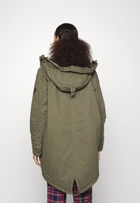 Giacca parka khaki con cappuccio, dotata di cordino, apertura posteriore e una toppa sul maniche sinistro. Modello che indossa pantaloni a quadri.