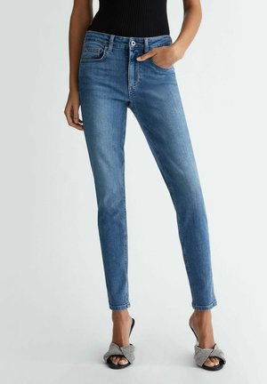 Vrouw draagt slim-fit blauwe spijkerbroek met een zwart mouwloos topje en open-toe zwarte hakken met geruite strikjes, staand tegen een effen achtergrond.
