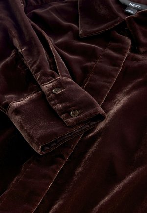 Ruskea samettipaita, jossa on kaulus ja napilliset kalvosimet. Siinä on pehmeä tekstuuri ja sileä viimeistely, jotka korostavat vaatteen rikasta väriä ja tyylikästä muotoilua.