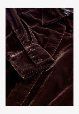 Ruskea samettipaita, jossa on kaulus ja napilliset kalvosimet. Siinä on pehmeä tekstuuri ja sileä viimeistely, jotka korostavat vaatteen rikasta väriä ja tyylikästä muotoilua.