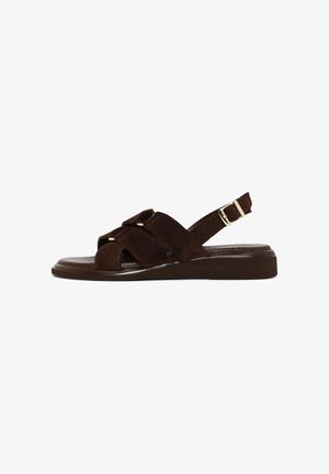 Sandalo piatto in suede marrone con cinturini incrociati e fibbia regolabile sul tallone, su suola testurizzata, mostrato di lato su sfondo bianco.
