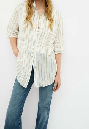 Femme portant une longue chemise blanche boutonnée avec des rayures verticales beige et un jean bleu à jambes larges, debout devant un fond blanc uni.
