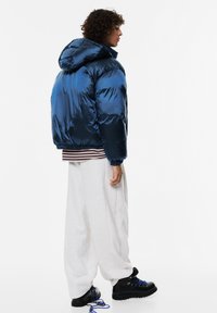 Niebieska kurtka puchowa z kapturem, błyszczący materiał, elastyczne mankiety, w zestawie z białymi spodniami dresowymi i czarnymi butami z niebieskimi sznurówkami.