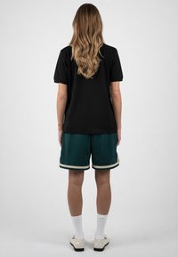 Personne aux longs cheveux blonds portant un t-shirt noir, un short vert avec une bordure beige, des chaussettes blanches et des chaussures blanches, debout de dos.