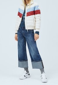 Prošívaná péřová bunda v béžové, modré a červené barvě se zipem vpředu; doplněná širokými džínami, šedými ponožkami a bílými teniskami.