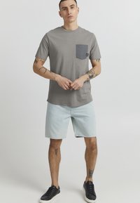 Gray cotton t-shirt with a dark gray pocket, paired with light denim shorts and black sneakers. Plain design, short sleeves, casual fit.