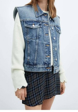 Kvinde iført en ærmeløs blå denimvest over en hvid striktrøje og et sort og blåt ternet skørt.