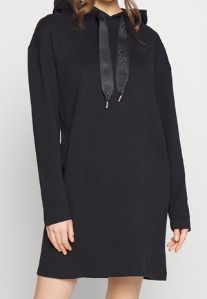 Kvinde iført en sort langærmet hættetrøjekjole med brede satin snore med tekst mod en ensfarvet, lys baggrund.