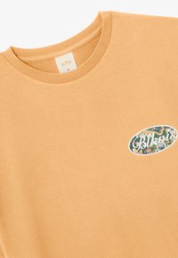 T-shirt en coton orange clair, col rond, avec un graphique ovale présentant un design multicolore complexe et du texte sur la partie gauche de la poitrine.