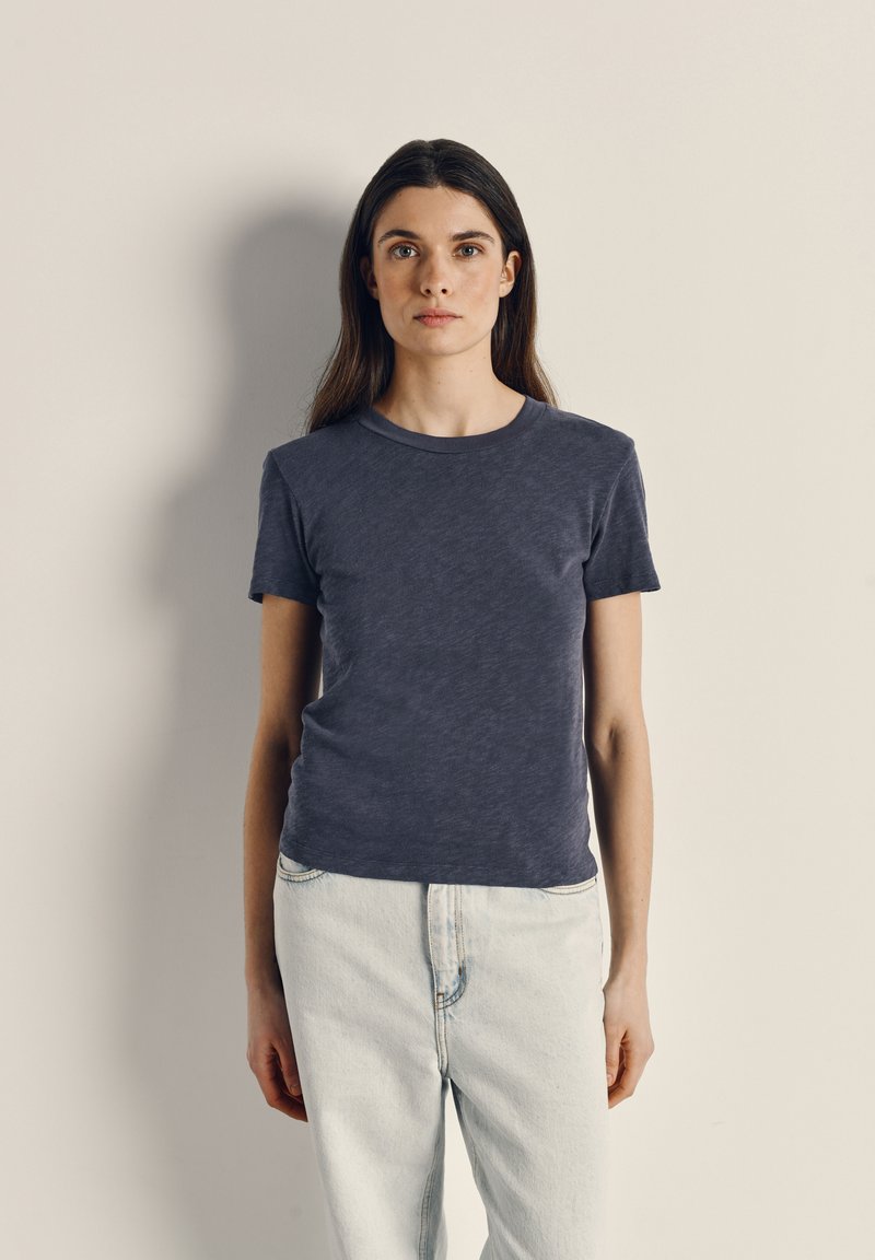 Young woman with long brown hair wearing a dark gray t-shirt and light blue jeans standing against a plain beige wall.