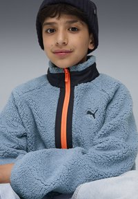 Ljusblå fleecejacka med hög krage, svart dragkedja och detaljer, har en framträdande orange dragkedja och svart logotyp på bröstet.