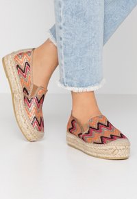 Fötter i färgglada espadriller med zigzag-mönster och tjocka flätade sulor, tillsammans med ljusblå jeans med fransig nederkant, på vit golv.