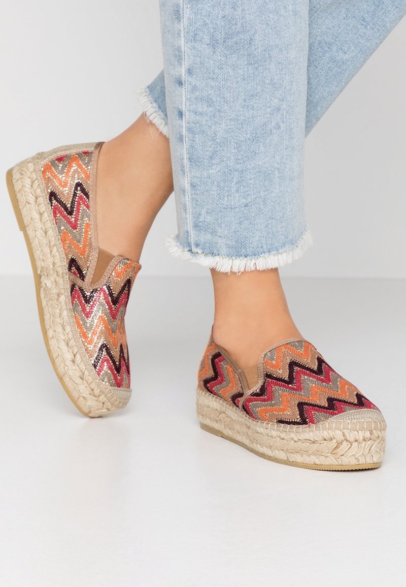 Fötter i färgglada espadriller med zigzag-mönster och tjocka flätade sulor, tillsammans med ljusblå jeans med fransig nederkant, på vit golv.