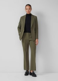 Vrouw in een olijfgroene, op maat gemaakte kostuum met een zwarte coltrui en zwarte instapschoenen, staande tegen een effen lichtgrijze muur.