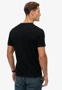 Camiseta de algodón negra con mangas cortas, cuello redondo y detalles de costuras en contraste. La parte trasera presenta un pequeño logo en la manga.