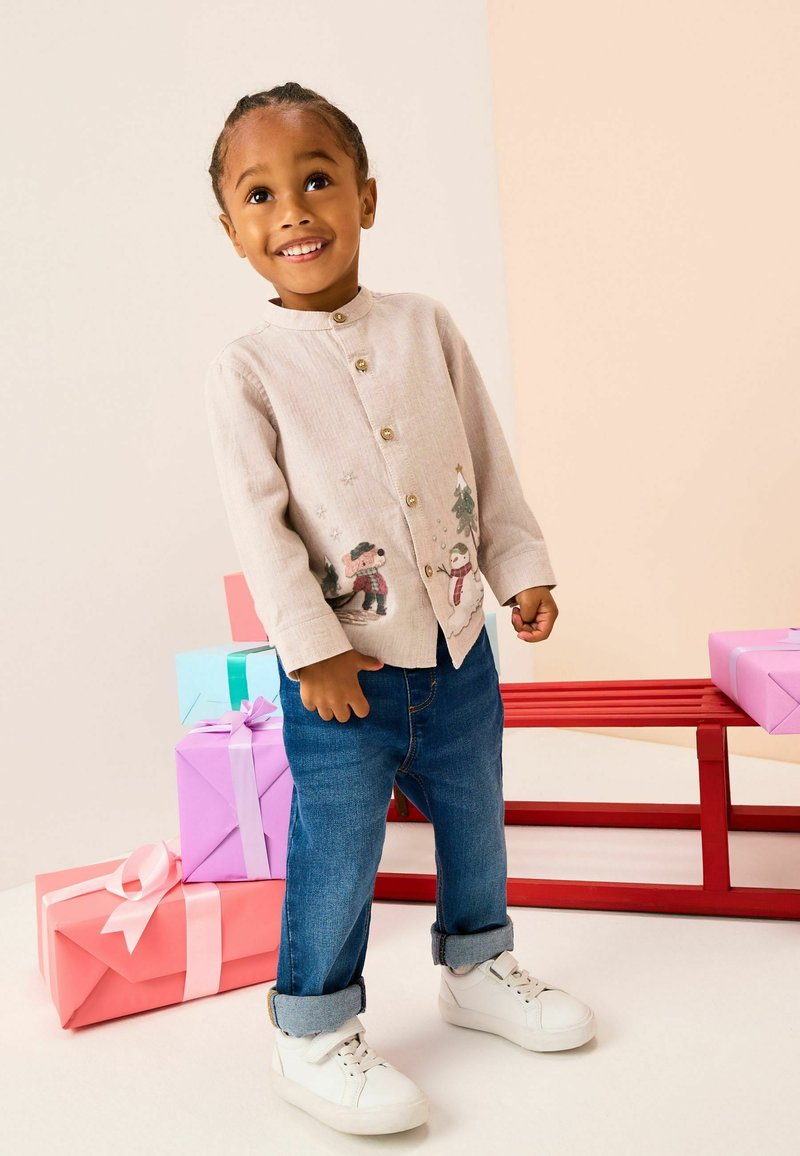 Helle beige Hemdbluse mit gesticktem Schneemann und Baum, kombiniert mit blauen Jeans und weißen Turnschuhen. Bunte Geschenkboxen im Hintergrund.