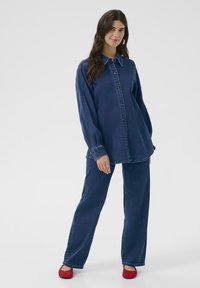 Denim blúz gombos elülső résszel, hegyes gallérral és hosszú ujjal. Széles lábú nadrággal párosítva, mindkettő sötétkék, piros cipővel kiegészítve.