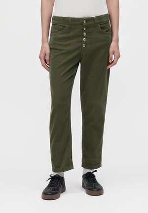 Personne portant un pantalon vert à boutons, des chaussettes blanches, des baskets noires et un haut blanc, les mains détendues le long du corps.