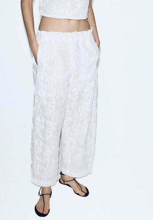 Weiße Hosen mit weitem Bein und floralem Muster, Hände in den Taschen, kombiniert mit schwarzen flachen Riemchensandalen und einem weißen Cropped-Top.