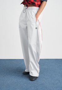 Jasnoszare, oversize spodnie z elastycznym paskiem w talii, bocznymi kieszeniami, czerwonymi bocznymi paskami i subtelną naszywką z logo. Do tego czarne buty wsuwane.