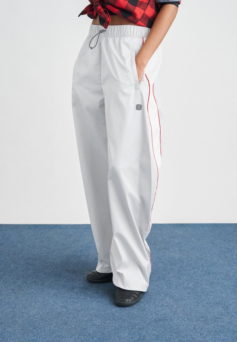 Jasnoszare, oversize spodnie z elastycznym paskiem w talii, bocznymi kieszeniami, czerwonymi bocznymi paskami i subtelną naszywką z logo. Do tego czarne buty wsuwane.