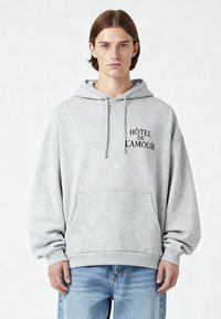 Szürke kapucnis pulóver elülső zsebbel, fekete „HÔTEL DE L'AMOUR” felirattal. Puha anyag, laza szabás és állítható húzózsinórral ellátott kapucni.