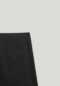 Pantalon noir à texture lisse, taille élastique, et une poche arrière unique avec une petite étiquette de marque. Design simple, sans motifs.