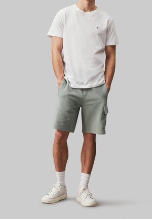 Hombre con camiseta blanca Calvin Klein, pantalones cortos cargo gris claro, calcetines blancos y zapatillas blancas, de pie con las manos en los bolsillos.