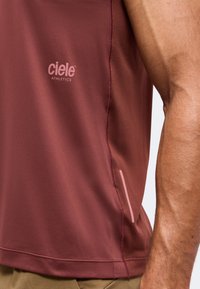 Débardeur sportif bordeaux avec le logo Ciele Athletics porté par une personne avec un bras visible et un pantalon beige.