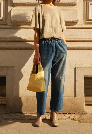 Béžové oversized tričko s velkým logem "M", doplněné volnými modrými džínovými kalhotami, taupe kotníkovými botami na podpatku a žlutou kabelkou.