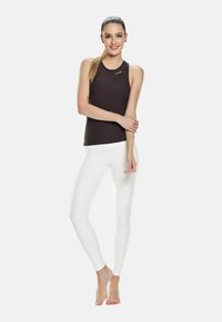 Kvinde, der står barfodet med en hestehale, iført en sort ærmeløs top og hvide tætsiddende leggins mod en ensfarvet baggrund.