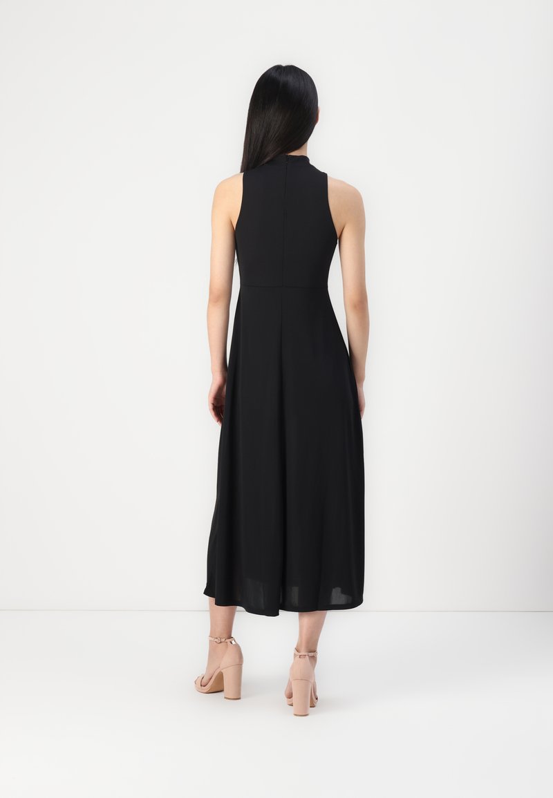 Schwarzes ärmelloses Maxikleid mit hohem Kragen, fließendem Stoff und tailliertem Oberteil. Dazu werden beigefarbene High Heels getragen.