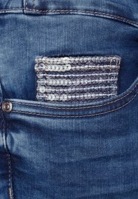 Jeans i denim med en bakficka prydd med silverpaljetter arrangerade i horisontella ränder och kontrasterande gula sömmar.