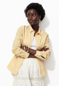 Chemise boutonnée à rayures jaunes et blanches avec des manches longues, portée par-dessus un débardeur blanc et une jupe blanche texturée avec des détails en œillets.